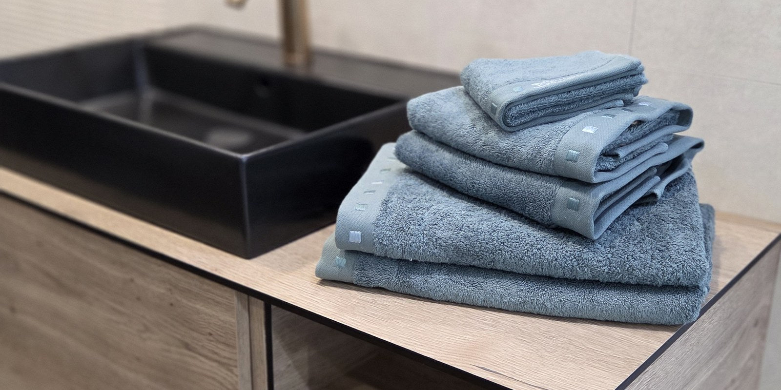 Three folded towel sizes stacked on a wooden bathroom vanity beside a black basin